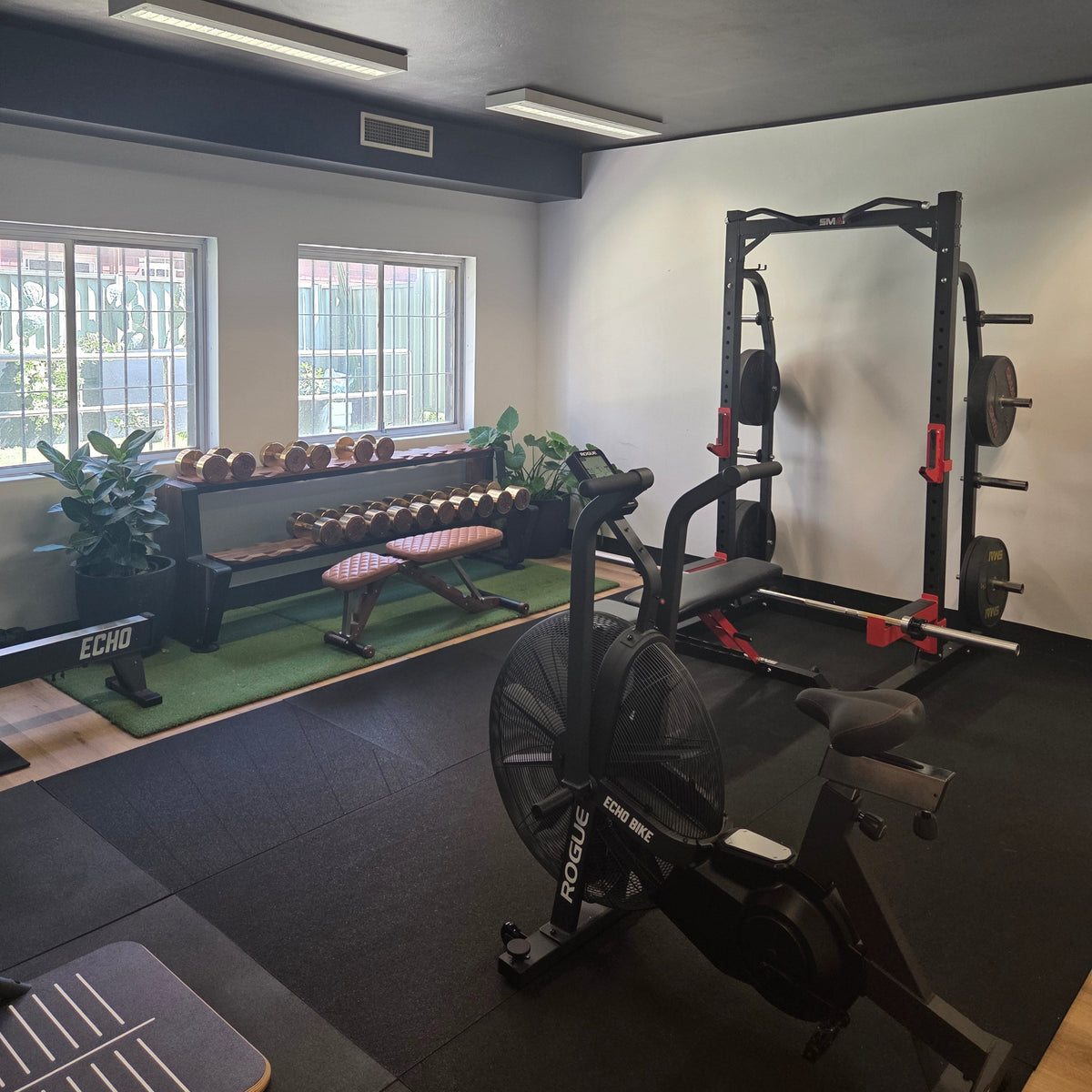 A home gym interior featuring a black Rogue fitness stationary bike in the foreground, a black power rack with weight plates on the right, and a weight bench with dumbbells on the left, set on black rubber flooring.